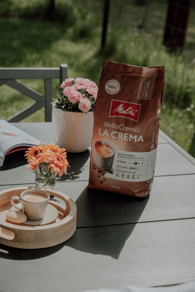 Kawa ziarnista Melitta BellaCrema LaCrema na stoliku wraz z filiżanką kawy, ciastkiem i kwiatkami.